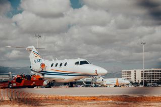 A private jet stationed on an airport tarmac with cloudy skies and mountains in the background.